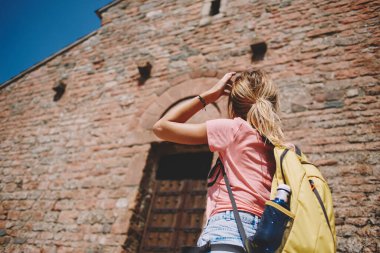 Sırt çantalı, saçlarına dokunan ve yaz aylarında ahşap kapılı eski taş binanın ön cephesine bakan kimliği belirsiz kadın gezginin arka planı.