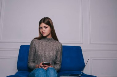 Akıllı telefonlu sıradan kıyafetler içinde düşünceli bir öğrenci kameraya bakıyor ve beyaz duvara karşı dizüstü bilgisayarla kanepede oturuyor.