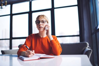 Ciddi bir uzaktan kumandalı kadın çalışanın masasına oturup telefon görüşmesi yapması ve çalışırken açık defterine notlar yazması.