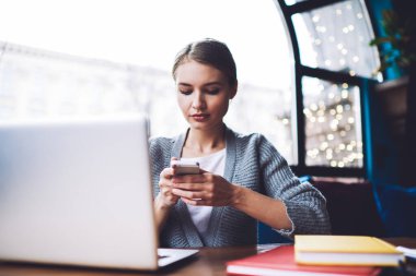 Gündüz vakti kafede dizüstü bilgisayar ve defterlerle masada otururken akıllı telefon kullanan düşünceli bir kadın.
