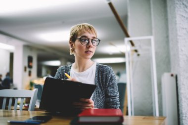 Gündelik giyim ve gözlük takan düşünceli kadın çalışma alanında uzaktan çalışırken not defterine bir şeyler yazıyor.