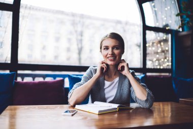Günlük kıyafetli, kulaklıklı, ahşap masada elinde defterle oturan ve gündüzleri rahat kafeye bakan tatmin olmuş bir kadın.