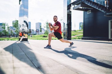 Şehir merkezindeki kaldırımlı yolda koşup kameraya bakmadan önce spor giyim kuşamında kaslı bir erkek sporcunun bacaklarını esnetirken yan görüntüsü.