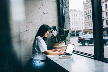 Günlük kıyafetlerle gülümseyen etnik bir kadının yan görünüşü kafede, masada, defterin yanında ve akıllı telefonun yanında uzaktan bilgisayarla çalışması.