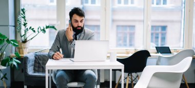 Ciddi sakallı girişimci masadaki dizüstü bilgisayarına bakar telefonda konuşur ve iş günlerinde not alır.