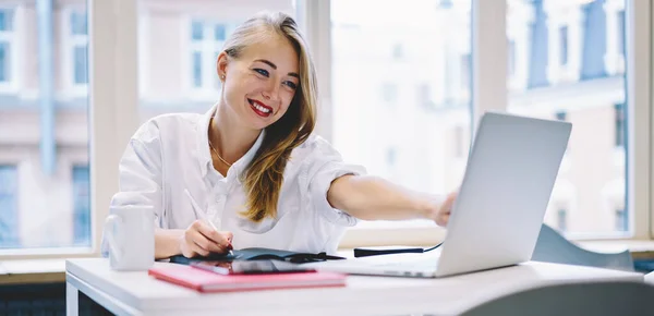 穿着白衬衫的年轻而积极的女性坐在桌旁 一边做笔记 一边看着现代办公室的笔记本电脑屏幕 — 图库照片