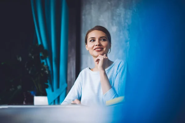 Happy brown haired female in casual clothes holding hand on chin and smiling while sitting at table and looking away