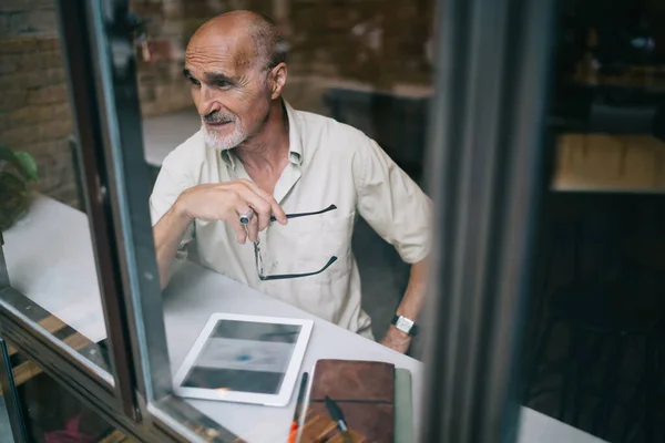 elderly male blogger with modern touch pad for browsing thinking about web information in cafe interior