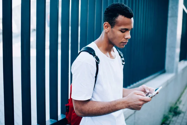 Side view of focused African American male with backpack browsing modern cellphone while standing near metal enclosure on street in city