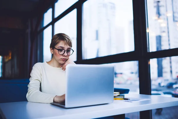 Concentrated female remote worker in casual clothes with short hair looking at screen of laptop while working on new project