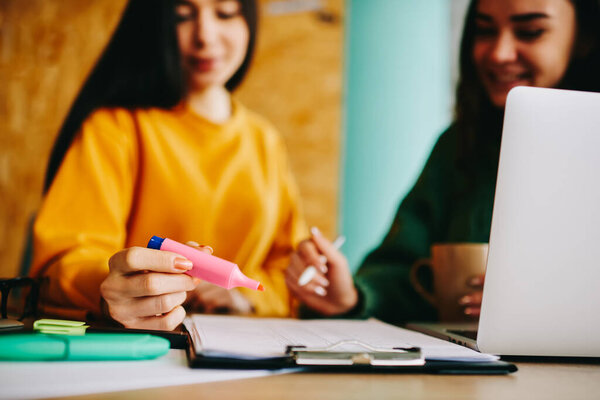 Crop young women in casual clothes looking down sitting at table with marker and pen writing notes on sheet of paper