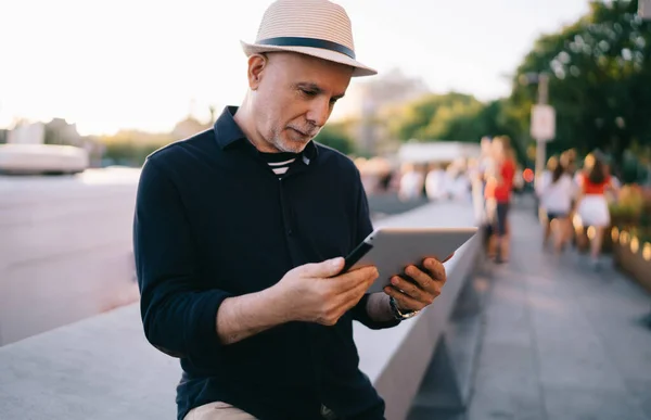 白种人男性老年游客在草帽网络社交媒体上通过现代触摸板阅读网站上的旅游建议 — 图库照片