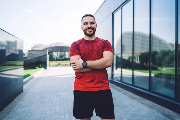 Positive male athlete in red tee shirt with wristwatch looking at camera and resting after workout while spending time in city