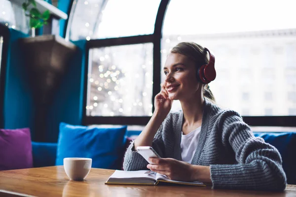 Satisfied woman in casual clothes listening to music on smartphone and looking away while taking notes in cozy cafe with cup of coffee