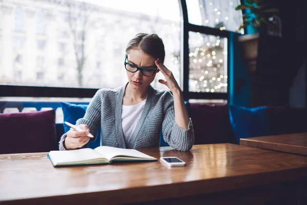 Frowning Female Student Eyeglasses Looking While Sitting Table Opened Notebook — Stockfoto