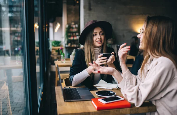Çalışan malzemelerle masada otururken ve birbirlerine bakarken bilgisayar konuşan ve el kol hareketi yapan modaya uygun giyinmiş genç kadınlar.