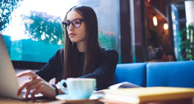 Kafe 'de çalışırken bilgisayar ekranına bakan günlük giysiler içinde yoğun kadın serbest çalışanın geniş açılı görüntüsü.