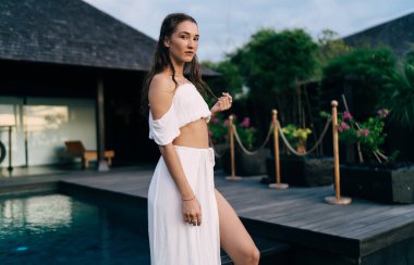 İyimser bayan gezgin, tatil sırasında yüzme havuzu yakınlarındaki tropik çiçeklerin bulanık manzarasına karşı verandada duran kameraya bakıyor.