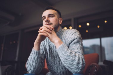 Concentrated man in grey sweater thoughtfully looking away while sitting in armchair and chilling in bright room decorated with lights