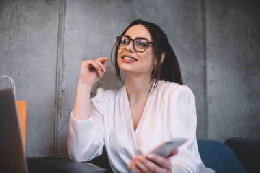 Modern çalışma ortamında dizüstü bilgisayarla masada otururken gülümseyen ve akıllı telefon tutan şık giyinen kendine güvenen kadın girişimci.