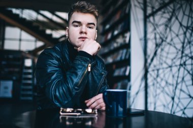 Deri ceketli ciddi bir genç adam modern kütüphanede elinde kupayla oturmuş çenesine dokunuyor ve dikkatlice kameraya bakıyor.