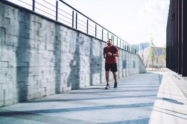 Tam boy genç sporcu kaldırım boyunca koşuyor ve kulaklıkları müzik için kullanıyor. Sabahları uzaklara bakıyor ve ısınıyor.