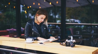 Modern kafeteryadaki masada oturan, elinde telefon, bardak içki ve fotoğraf makinesiyle ilginç edebiyat kitapları okuyan konsantre genç bir kadın.