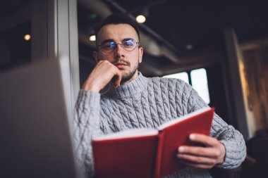Ciddi sakallı, sıcak süveter ve gözlüklü bir erkek dizüstü bilgisayarla çalışıyor ve modern iş yerinde kitapla otururken kameraya bakıyor.