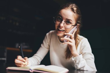 Beyaz kazaklı ve gözlüklü genç bir kadın cep telefonuyla yeni projeyi tartışıyor ve masada oturan planlamacıya not alıyor.