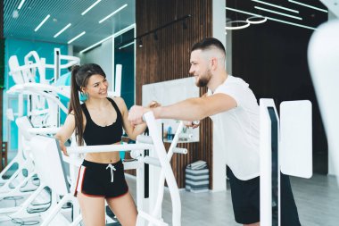 Modern spor salonunda sporcu eğitimiyle sohbet ederken, spor makinesindeki dijital monitör ekranına dokunarak gülümseyen formda bir sporcu kadın.