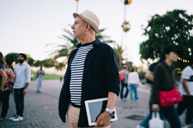 Emeklilik günlerinde modaya uygun giyinmiş beyaz erkek blogcu, elinde modern bir dokunmatik ped, yürüme saatinde başka yere bakıyor.