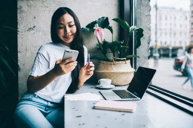 Gündelik giyinen genç Asyalı pozitif bir kız öğrenci kafede oturuyor ve ödevlerini yaparken mesaj atıyor ve not alıyor.
