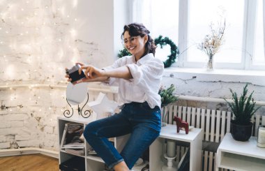 Neşeli kadın gülümsüyor ve eski moda kamerayla kendi portresini çekiyor. Sıcak bir odanın penceresinin yanında duruyor ve duvarında parlayan ışıklar var.