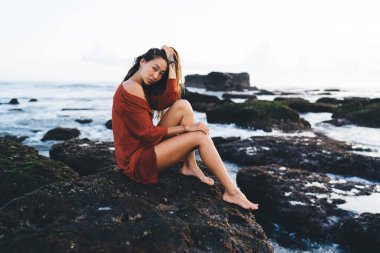 Bali 'nin kayalık sahillerinde oturmuş kameraya bakıp saçlarına dokunan mini etek giysili genç bir kadının yan görüntüsü.