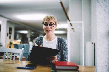Genç gülümseyen bayan çalışan, kameraya bakıyor. Masada not defterleri ve akıllı telefon notlarıyla oturuyor. Çalışma saatleri boyunca pano kullanarak notlar yazıyor.