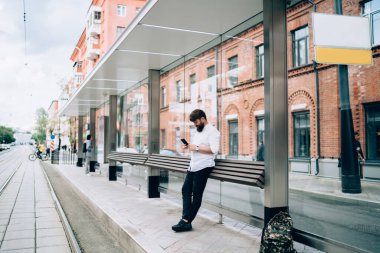 Sırt çantalı yetişkin bir adam tramvay istasyonunda duruyor ve bulutlu bir günde boş şehir caddesinde tuğladan binalara karşı akıllı telefonlara bakıyor.