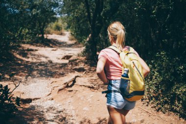 Sarı sırt çantası taşıyan, yazın güneşli bir günde tozlu yolda yürüyüşe çıkan kimliği belirsiz genç bir kadının arka görüntüsü.