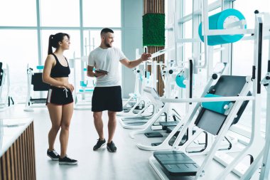 Sportif giyinen sporcu kadın ve kas koçu modern fitness makineleri boyunca spor salonunda yürürken antrenman için ekipman seçiyor.