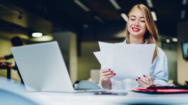 Masadaki modern kafede oturan gülümseyen kadın beyaz gömlek giyerken bilgisayar ve kağıtları uzaktan çalışma için kullanıyor.