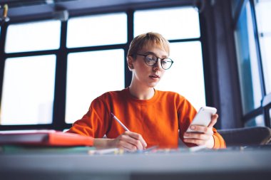 Turuncu kazaklı genç kız öğrenci ve gözlüklü ev ödevlerini yaparken cep telefonunu karıştırıyor.