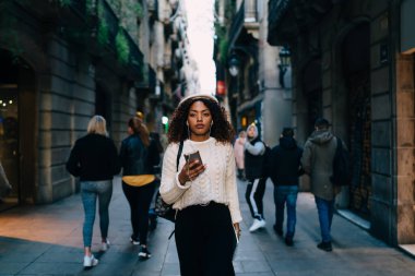 Havalı Afro-Amerikan kadın, günlük kıyafetler ve bereyle dar bir sokakta yürüyor ve akıllı telefonlara bakarak müzik dinliyor.