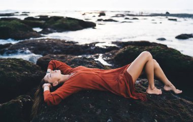 Çıplak ayaklı çekici bir kadın saçlarına dokunuyor ve Bali 'de eğlenirken kayalık sahilde vakit geçiriyor.