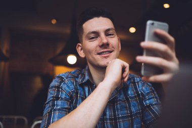 Gündelik kıyafetlerle gezen, akıllı telefonlara bakan ve elleri çenesinde oturmuş gülümseyen ve kameraya bakan neşeli genç adam.