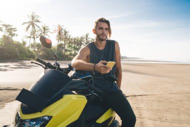 Ciddi genç adam, günlük giysiler içinde güneşli bir günde deniz kenarında bisiklete biniyor ve kameraya bakarken cep telefonuna bakıyor.