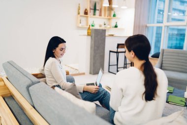 Bilgisayarlı bir kadının, geniş oturma odasındaki modern bir kanepede otururken ekranda kız arkadaşıyla sohbet edişinin yan görüntüsü.