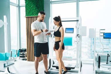 Spor giyim sektöründe tıraş edilmemiş genç bir eğitmen hafif spor salonundaki elektronik makine ve pencerelerin yanında konuşurken gülümseyen bir kadına tablet gösteriyor.