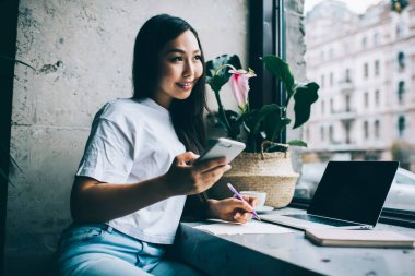 Bilgisayarda ev ödevi yapan mutlu Asyalı genç kız öğrenci ve kafede notlar alıp akıllı telefondan sohbet ediyor ve gözlerini kaçırıyor.