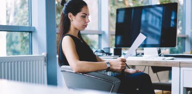Şık giyinmiş, ofis masasında dizüstü bilgisayarla oturan ve gündüzleri evrak okuyan konsantre iş kadınının yan görüntüsü.