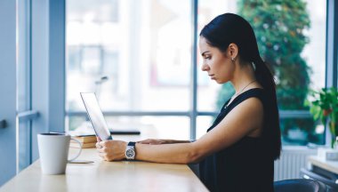 Odaklanmış, siyah saçlı, netbook üzerinde çalışan, masada oturup, çağdaş iş yerinde ekrana bakan kadın serbest yazar.