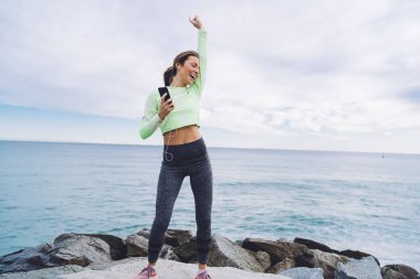 Spor kıyafetli, taşların üzerinde dans eden ve deniz kenarında akıllı telefon ve kulaklık kullanan mutlu genç kadın.